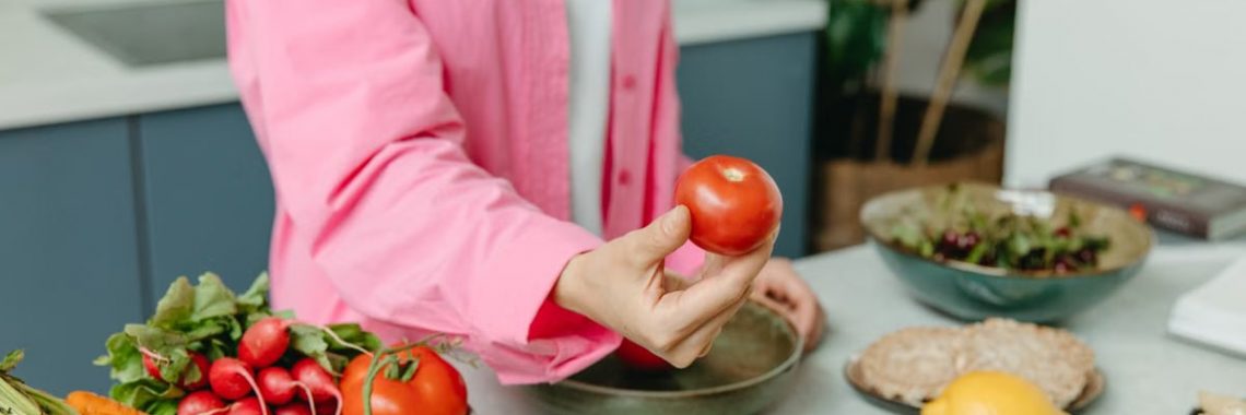 Person in pink overshirt holding a fresh tomato with various organic vegetables on a countertop.