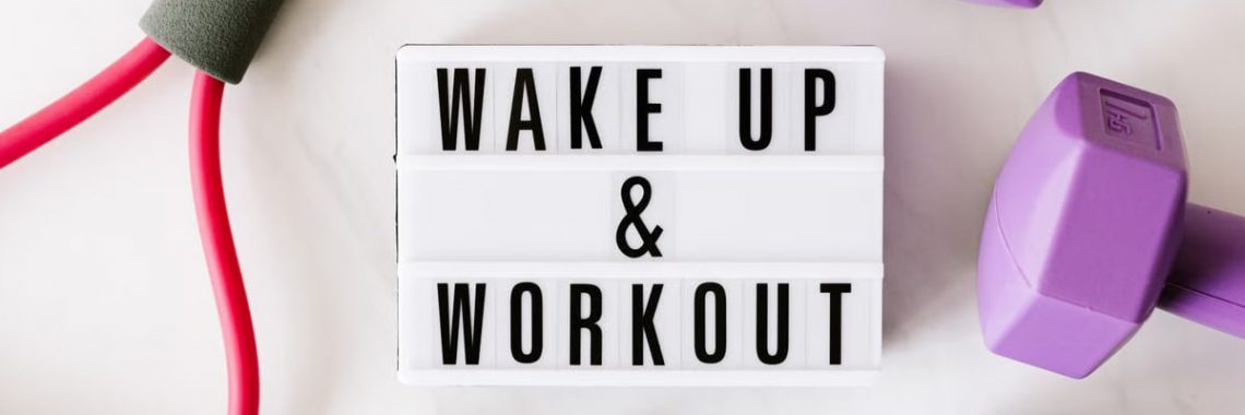 Top view closeup composition of wake up and workout inscription on light box surface among expanders and dumbbells and massage ball on white background